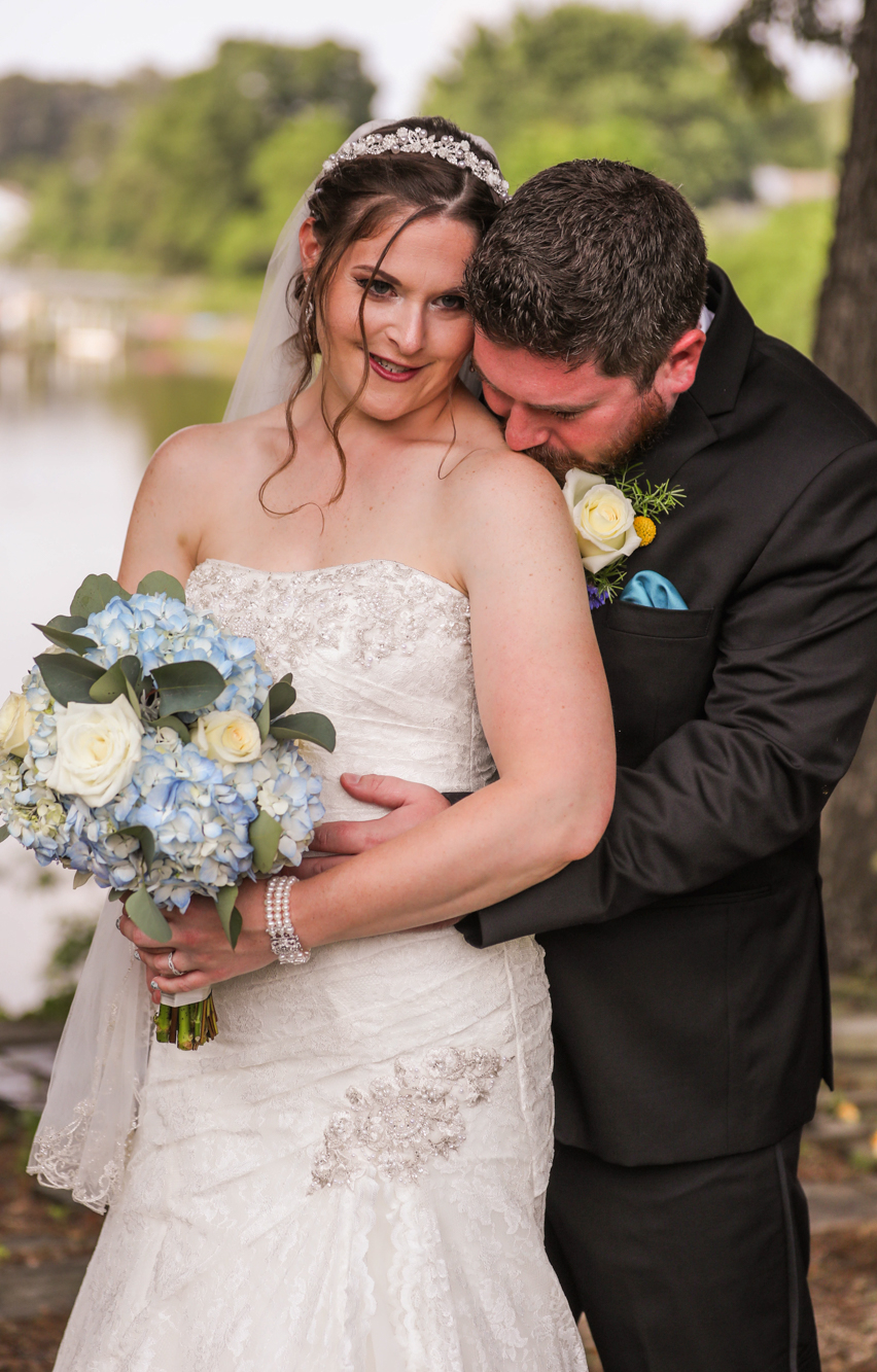 Groom nuzzling bride holding a blue hydrangea bouquet by the lake